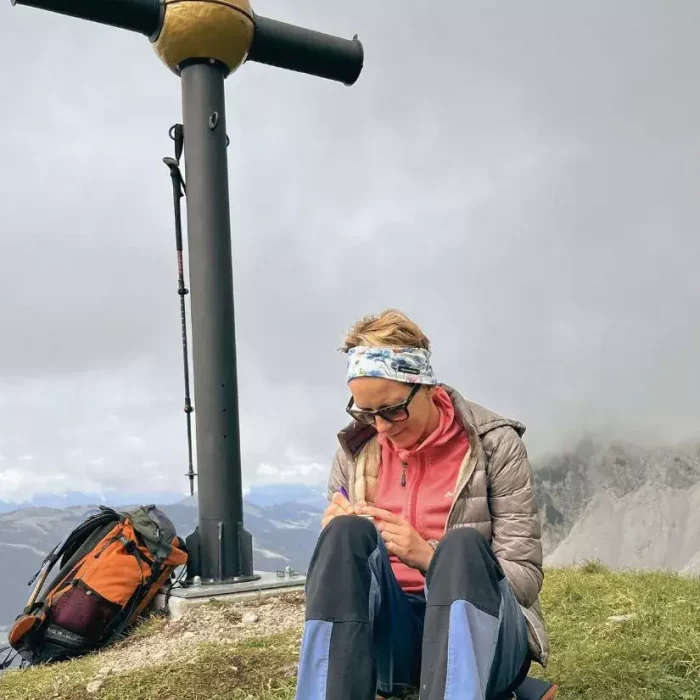 Nahaufnahme einer Person, die am Gipfelkreuz sitzt und auf einem Zettel / Notizblock etwas notiert.